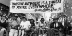 Black and white photo of several people in the middle of a city street, in wheelchairs. More are coming up behind them and two people are carrying a banner reading "'Injustice Anywhere Is a Threat to Justice Everywhere' -- Martin Luther King, Jr."
