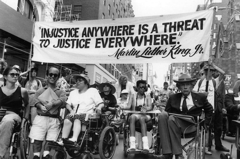 Black and white photo of several people in the middle of a city street, in wheelchairs. More are coming up behind them and two people are carrying a banner reading "'Injustice Anywhere Is a Threat to Justice Everywhere' -- Martin Luther King, Jr."