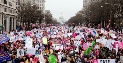 A crowd of people, many with feminist protest signs, on Pennsylvania Avenue in Washington, DC. The Capitol Building is visible in the center background.