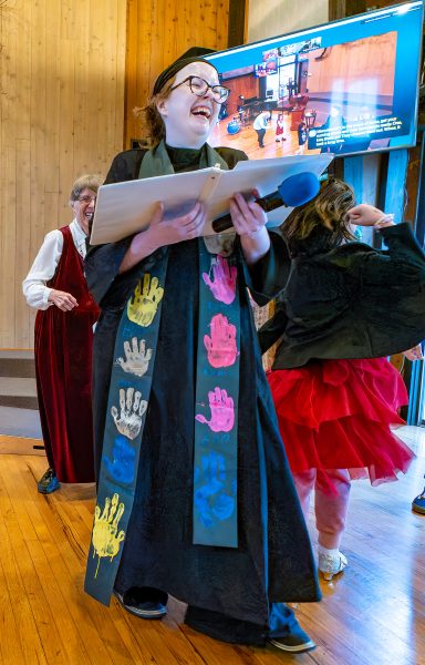 Young white, female minister, wearing a black robe and a stole patterned with children's handprints, laughing in the congregation's Main Hall